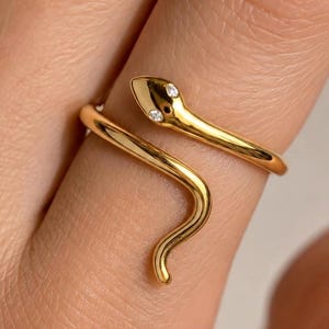 May include: A gold-toned wrap ring shaped like a snake, featuring a smooth, polished finish and two small, clear, round-cut stones set as eyes on the snake's head. The band curves around the finger in a continuous, open-ended coil design.