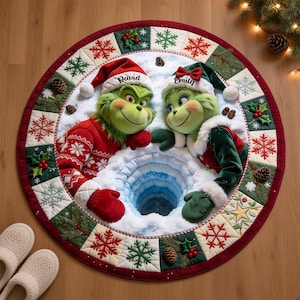 Könnte beinhalten: Ein runder Weihnachtsbaumteppich mit zwei grünen Figuren in Weihnachtsmannmützen, eine in einem roten Pullover und eine in einem grünen Samtgewand. Die Namen David und Emily sind auf die Mützen gestickt. Der Rand zeigt ein gestepptes Muster aus Schneeflocken, Tannenzapfen und Stechpalmenbeeren um ein zentrales, verschneites Lochdesign.