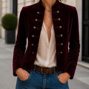 Peut inclure: Une veste en velours bordeaux profond dotée d'un col montant, de manches longues et d'une fermeture à double boutonnage avec des boutons métalliques argentés. La veste est portée sur une chemise blanche, un jean bleu et une ceinture en cuir marron.