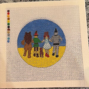 May include: A needlepoint canvas featuring a circular design of four figures and a small dog walking on a yellow path against a blue sky. A vertical column of colored thread squares is visible on the left side of the white grid fabric.
