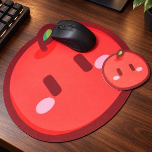 May include: A large, round, red mouse pad featuring a stylized apple character with a small green leaf, dark red rectangular eyes, and pink oval cheeks. A smaller, matching circular coaster sits to the right, and a black computer mouse rests on top.