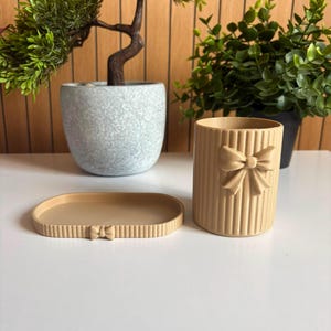 May include: A beige desk organiser set consisting of a cylindrical pen holder and an oval tray. Both items feature a vertical ribbed texture and a small, moulded bow detail on the front. The pen holder is upright, while the tray is flat.