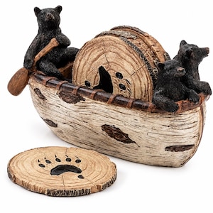May include: A decorative coaster set featuring a holder shaped like a birch bark canoe with three black bear figures. The set includes circular coasters designed to resemble wood slices, each featuring a black bear paw print in the center.