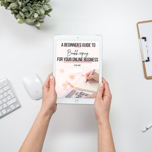 May include: A white tablet displaying a pink and white ebook cover with the title "A Beginner's Guide to Bookkeeping for Your Online Business". The cover features a hand writing in a notebook with a calculator.