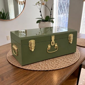 May include: A rectangular olive green metal storage trunk with a glossy finish. The box features gold-toned metal hardware, including two side handles, two front latches, and a central locking mechanism with a hinged lid design.