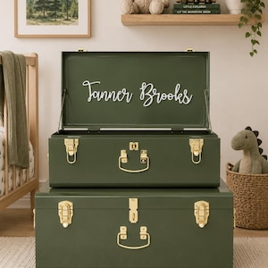 May include: A set of two stacked olive green metal storage trunks with gold-colored latches and handles. The top trunk is open, revealing the white script text Tanner Brooks on the inside of the lid. Both trunks feature a smooth, solid finish.