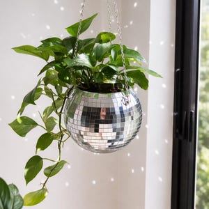 May include: A spherical disco ball planter covered in small, reflective silver mirror tiles. The planter is suspended by a silver metal chain and contains a lush green pothos plant with trailing vines that spill over the sides of the container.