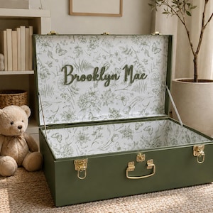 May include: An olive green storage trunk with gold-toned metal latches and a handle. The interior is lined with white paper featuring a green botanical and butterfly print. The name Brooklyn Mae is displayed in raised green script on the lid.