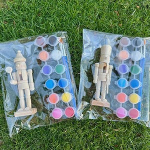 Puede incluir: Dos bolsas de plástico transparente que contienen cada una una figura de cascanueces de madera sin pintar, una tira de diez pequeños botes de pintura de varios colores y un pincel. Los cascanueces son de color beige claro con brazos articulados.