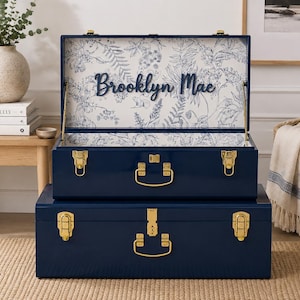 May include: A set of two navy blue storage trunks with gold-toned metal latches and handles. The top trunk is open, revealing an interior lining featuring a white and blue botanical floral pattern with the name Brooklyn Mae in dark blue script.