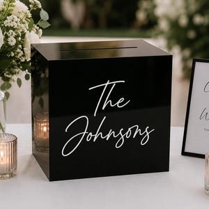 May include: A square black acrylic box with a horizontal slot on top for collecting cards. The front of the box features the white script text "The Johnsons" printed in the center. The box has a smooth, reflective surface and a clean design.