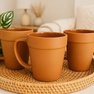 May include: Three terracotta-colored ceramic mugs with a tapered shape and a wide rim, resembling the appearance of flower pots. The mugs feature a smooth matte finish and a curved handle, arranged together on a round, light-colored woven rattan tray.