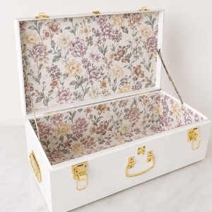 May include: A white rectangular storage chest with gold-toned metal latches and handles. The interior is lined with a floral pattern featuring purple, yellow, and green flowers on a cream background. The lid is open and held by metal hinges.