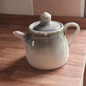 Może przedstawiać: Ceramiczny czajniczek o zaokrąglonym korpusie i dopasowanej pokrywce ze spiczastym uchwytem. Czajniczek posiada gradientowe szkliwo, przechodzące od kremowej bieli u podstawy do przygaszonej szałwiowej zieleni i ciemnej szarości przy górnej krawędzi.