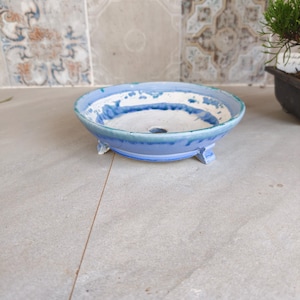 May include: A shallow, round ceramic bonsai pot featuring a blue and white glazed finish with a mottled, textured appearance. The pot is supported by three small feet and includes a central drainage hole at the bottom of the basin.