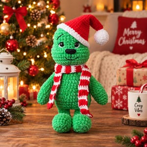 Puede incluir: Un peluche de ganchillo verde con forma de criatura y nariz negra, que lleva un gorro de Papá Noel rojo y blanco y una bufanda a rayas rojas y blancas. El juguete tiene una cabeza redondeada, dos brazos y dos piernas de hilo texturizado.