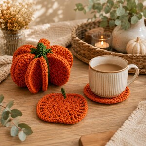 May include: A set of orange crochet pumpkin-shaped coasters, including a three-dimensional pumpkin storage piece with a green stem and several flat, circular pumpkin-themed coasters, one of which supports a ribbed ceramic mug filled with coffee.