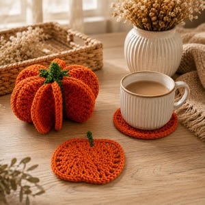May include: A three-dimensional crocheted orange pumpkin with a green stem, a flat crocheted pumpkin-shaped coaster, and a white ceramic mug with a ribbed texture resting on a matching orange crocheted circular coaster.