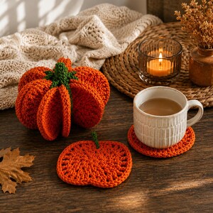 May include: A crocheted orange pumpkin with a green stem, a flat crocheted pumpkin-shaped coaster, and a white ceramic mug resting on a matching orange crocheted coaster. The items are arranged on a dark wooden surface with a dried leaf nearby.