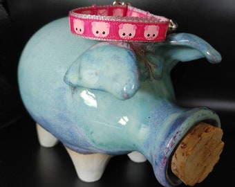 May include: A pink and white pig-themed collar with a silver buckle and a bell. The collar is sitting on top of a blue and white ceramic piggy bank.