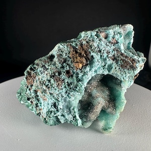 Peut inclure: Un spécimen minéral brut présentant une structure cristalline vert turquoise vif avec des inclusions poreuses, brun foncé et aux tons métalliques. La roche a une texture de surface irrégulière et dentelée avec un centre creux rempli de fins cristaux.