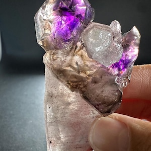 Può includere: Un gruppo di cristalli grezzi caratterizzato da una grande base traslucida con inclusioni scure, sormontata da diverse terminazioni appuntite e affilate. Le sezioni superiori mostrano vivaci tonalità viola intenso di ametista.