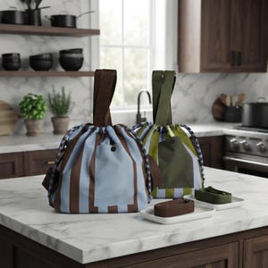 Puede incluir: Dos bolsas con cierre de cordón y asas superiores. Una bolsa presenta rayas verticales azul claro y marrón con un bolsillo frontal azul claro liso. La otra bolsa tiene rayas verticales verdes y blancas con un bolsillo frontal verde liso. Dos correas pequeñas a juego descansan sobre bandejas blancas.
