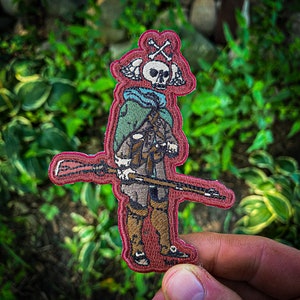 May include: A red, white, and blue embroidered patch of a skeleton holding a rifle. The skeleton is wearing a green and brown jacket and has a skull with crossed bones on its head.