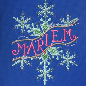 以下が含まれることがあります： 青い背景に、雪の結晶のモチーフとピンクの文字で「MARIEM」と書かれた刺繍のデザインです。