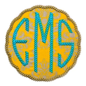 May include: A round, yellow fabric monogram with a gold rope border. The monogram features the letters "EMS" in a turquoise rope-like design. The background has a subtle, repeating pattern in a lighter shade of yellow.