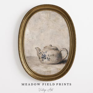 May include: An oval-framed wall art piece featuring a painting of a cream-colored ceramic teapot with blue floral patterns. The teapot sits on a textured surface against a neutral, mottled background. The frame is ornate, metallic gold, and oval.