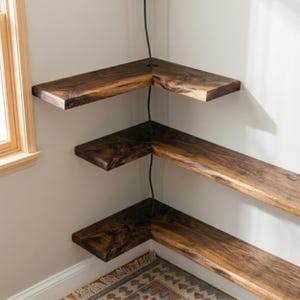 Op de afbeelding: Drie L-vormige hoekplanken van donkerbruin hout met een zichtbare houtnerf. Elke plank heeft een ronde uitsparing bij de hoek, waar een zwart snoer verticaal doorheen loopt en de drie niveaus met elkaar verbindt.