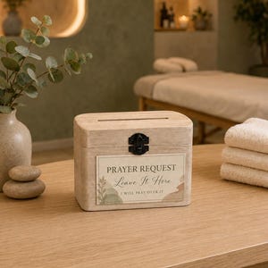 May include: A light-colored wooden box with a rectangular slot on top and a black metal latch on the front. A rectangular label is attached to the front, featuring the text PRAYER REQUEST Leave It Here I WILL PRAY OVER IT with botanical art.