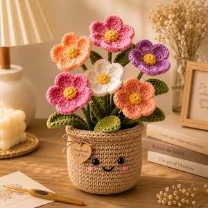 May include: A crochet flower arrangement featuring pink, orange, white, and purple blossoms with yellow centers and green leaves. The flowers are held in a tan, cylindrical crochet pot with a smiling face and a heart-shaped tag reading Handmade with love.