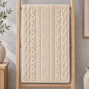 May include: A cream-coloured knit throw blanket featuring a complex arrangement of vertical cable, diamond, and bobble stitch patterns. The rectangular textile displays a symmetrical design with textured columns running the full length of the piece.