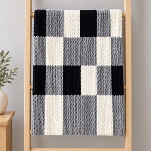 Könnte beinhalten: Eine grob gestrickte Wohndecke mit einem sich wiederholenden Karomuster aus abwechselnden schwarzen, weißen und grauen Blöcken. Die Decke besteht aus dickem, weichem Garn mit einer sichtbaren, strukturierten Maschenoptik auf der gesamten Fläche.