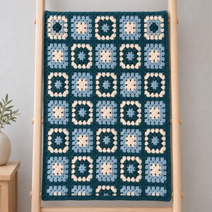 Puede incluir: Una manta de ganchillo rectangular con un patrón repetitivo de cuadrados de la abuela en tonos azul cerceta oscuro, azul claro y crema. Los cuadrados están unidos por un borde azul cerceta oscuro, creando una cuadrícula estructurada.