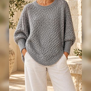 Puede incluir: Un suéter de punto gris con un patrón de diamantes texturizado, cuello redondo y mangas largas con puños acanalados anchos. El suéter tiene un dobladillo curvo y se combina con pantalones de color claro, creando un aspecto casual y superpuesto.