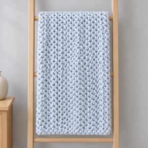 Könnte beinhalten: Eine hellblaue, grob gestrickte Decke mit einer dicken, strukturierten Garnkonstruktion und einem gleichmäßigen, sich wiederholenden Strickmuster. Die Decke hängt über einer Holzleiter und zeigt ihr weiches, voluminöses und einheitliches Gewebe.