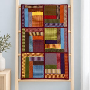 Può includere: Una coperta in maglia rettangolare caratterizzata da un design patchwork composto da sei grandi quadrati. Ogni quadrato è formato da piccoli segmenti rettangolari multicolori nelle tonalità del blu, giallo, arancione, verde, viola e rosso, bordati da una finitura rosso scuro.
