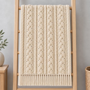 May include: A cream-colored knitted throw blanket featuring vertical cable knit patterns and textured columns. The blanket is finished with a row of long, straight fringe along the bottom edge, creating a uniform and structured appearance throughout.