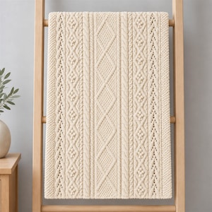 Peut inclure: Un jeté de lit en tricot de couleur crème présentant un motif complexe de torsades verticales, de losanges en treillis et de textures en relief. Le textile est rectangulaire avec un tissage dense, uniforme et cohérent sur toute la surface.