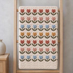 May include: A cream-colored crochet blanket featuring a repeating pattern of raised, textured tulips in rows of terracotta, dusty pink, light blue, and mustard yellow, each with dark green stems and leaves, arranged in a grid on a textured background.