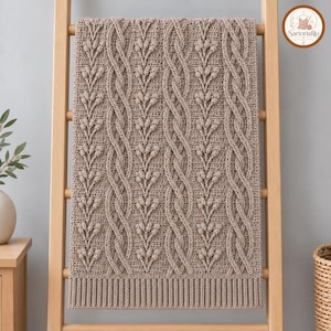May include: A beige crochet blanket featuring a textured pattern of vertical cable stitches and raised bobble stitch leaf motifs. The blanket has a ribbed border at the bottom edge and is draped over a light-colored wooden ladder frame.