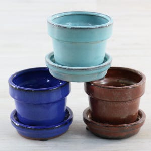 May include: Three small ceramic plant pots with matching saucers, arranged in a stack. One pot is light blue, one is deep royal blue, and one is a warm reddish-brown. Each pot features a smooth, glossy finish with a dark, defined rim.