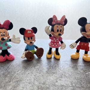 Peut inclure: Un ensemble de quatre figurines de personnages en plastique. La première est une souris en robe à pois bleu sarcelle avec un nœud rose. La deuxième porte une casquette rouge avec un M, un haut bleu et un pantalon vert. La troisième porte une robe à pois rose et un nœud. La quatrième porte un short rouge et des chaussures jaunes.