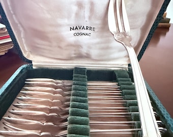 Op de afbeelding: Een set zilverkleurige metalen vorken in een donkergroene, gestructureerde presentatiedoos. De binnenkant van het deksel is bekleed met crèmekleurige stof met de tekst NAVARRE COGNAC. Eén vork staat rechtop tegen het deksel aan.