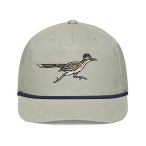 May include: A light gray baseball cap featuring a graphic illustration of a roadrunner bird in motion. The cap has a structured crown, a curved brim, and a dark navy blue rope detail running horizontally across the base of the front panel.