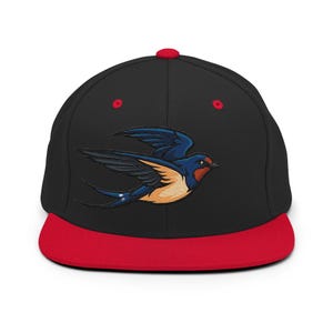 May include: A black snapback cap featuring a red brim and a red top button. The front panel displays an embroidered graphic of a flying swallow bird with dark blue wings, a reddish-brown throat, and a light tan underbelly against the black fabric.