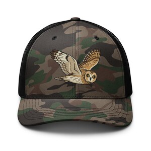 May include: A camouflage trucker hat featuring a black mesh back and a front panel with a printed illustration of a brown and tan owl in flight. The cap has a curved brim and a dark green, brown, and black camouflage pattern across the front.